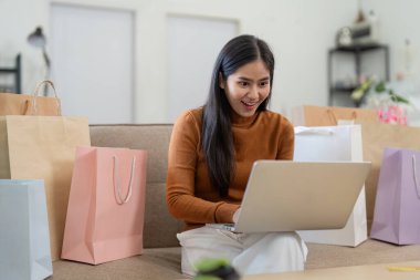 Bir kadın internetten bilgisayarını ve kredi kartını kullanarak alışveriş yapıyor. Etrafı alışveriş torbalarıyla çevrili. Modern ve rahat bir oturma odasında..