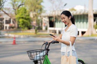 Genç bir kadın telefonunu yeşil bisikletinin yanında kullanırken gülümsüyor, modern ve sürdürülebilir bir iş seyahatini temsil ediyor..