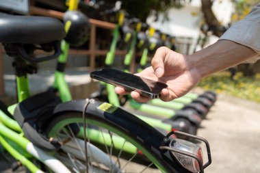 Bir banliyö çalışanı, bir bisiklet paylaşım programından yeşil bir bisikleti açmak veya kiralamak için akıllı telefon kullanır, modern ulaşım çözümlerinde teknolojinin rolünü vurgular.