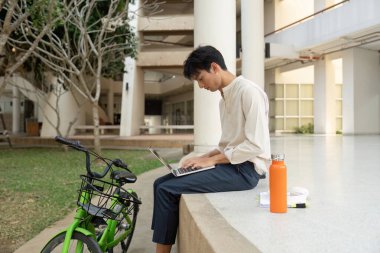 A young man sits comfortably working on his laptop in a lush urban environment, merging work and sustainable living through cycling. clipart