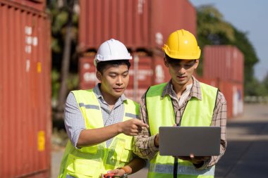 Güvenlik donanımlı iki genç mühendis dışarıda lojistik stratejilerini tartışırken bir laptopta veri analiz ediyorlar..