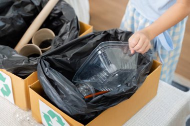 Bir çocuk geri dönüşüm projesine katılıyor, konteynırları dikkatlice belirlenmiş çöp kutularına yerleştiriyor, sorumlu atıkların atılmasının önemini öğreniyor.
