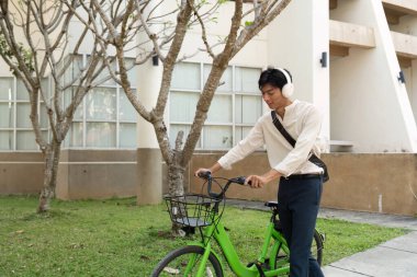 Kulaklık takan genç bir adam yeşil bisikletinin yanında yürüyor, kentsel ortamlarda sürdürülebilir ulaşımın önemini vurguluyor, sağlıklı bir yaşam tarzını teşvik ediyor..