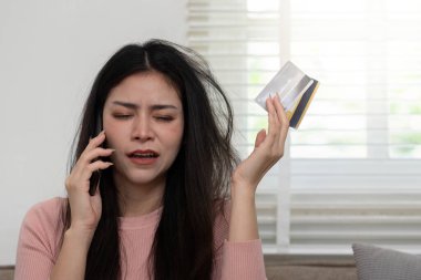 Bir kadın elinde kredi kartıyla telefonla konuşuyor. Konforlu evinde mali zorluklarla boğuşuyor..