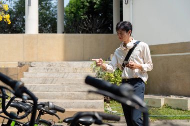 Bir birey bisiklet parkının yanında duruyor, hızlı, sürdürülebilir bir yolculuk için bisiklet bulmak için akıllı telefon kullanıyor, teknolojinin ve yeşil ulaşımın füzyonunu gösteriyor..