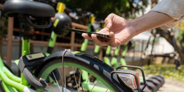 Bir kullanıcı elektrik bisikletine binmeye hazırlanırken telefonunu kontrol eder, yeşil bir şehirde sürdürülebilir ulaşım çözümlerini teşvik eder.