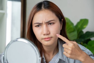 Genç bir kadın yansımasını inceliyor, derisi hakkında endişelerini dile getiriyor. Daha açık bir ten rengine ulaşabilmek için cilt bakımına ihtiyacı olduğunu düşünüyor..