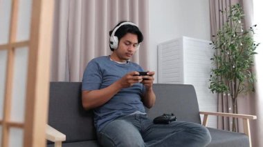 Oturma odasındaki kanepede oturan Asyalı genç bir çocuk, boş zamanlarında akıllı telefonuyla online oyun oynarken tamamen odaklanmış..