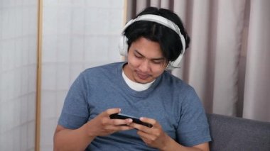 Oturma odasındaki kanepede oturan Asyalı genç bir çocuk, boş zamanlarında akıllı telefonuyla online oyun oynarken tamamen odaklanmış..