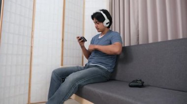 Oturma odasındaki kanepede oturan Asyalı genç bir çocuk, boş zamanlarında akıllı telefonuyla online oyun oynarken tamamen odaklanmış..