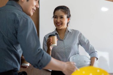 Bir mühendis ve meslektaşı, planlar ve parlak bir ofis ortamı içinde bir sonraki projelerini planlarken kahve içerken kısa bir anı paylaşıyorlar. Bu sahnede takım çalışması ve profesyonellik var..