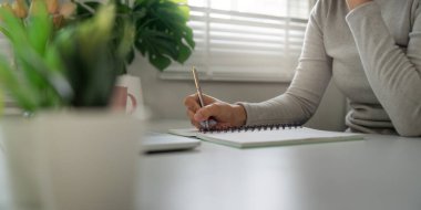 Dikkatle odaklanan bir birey, masasındaki bir deftere, bitkiler ve ışıkla çevrili bir şekilde, ev ortamında huzurlu bir çalışma sergilediğini yazar..