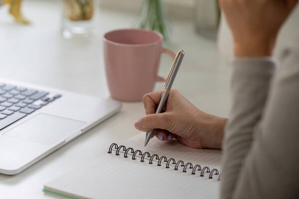 Сосредоточившись пристально, женщина пишет в своем блокноте рядом с ноутбуком, демонстрируя баланс письма и цифровой работы, потягивая кофе.