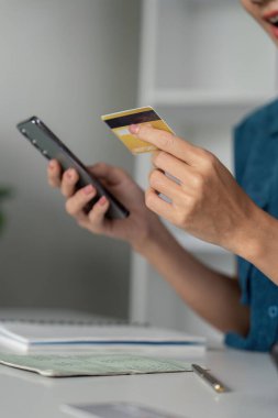 Bir kadın akıllı telefon alışverişi yapıyor, elinde bir kredi kartı tutuyor ve bütçesini not alıyor, günlük hayatta dijital alımların büyüdüğünü gösteriyor..