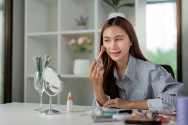 Bir kadın makyaj uygulamasına odaklanır, ustalıkla aynanın önünde fırçayla allık sürer. Huzurlu ortam onun güzellik rutinini arttırıyor..