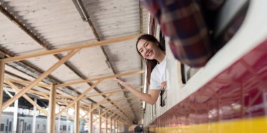 Neşeli bir gezgin, yeni bir yolculuğa hazır olduğunu belirterek, bir tren penceresinden dışarı bakarken heyecanını ifade eder. Hareketli istasyon seyahat atmosferine ekleniyor..