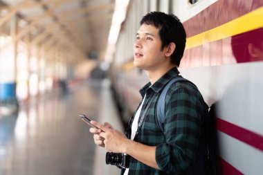 Genç bir gezgin, tren istasyonunun duvarına yaslanır, beklentiyle yukarıya bakar, her macerayla gelen bekleyiş anlarını gösterir..