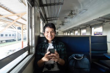 Gülümseyen genç bir adam trende rahat bir şekilde oturur, seyahat ve teknolojinin heyecanını somutlaştıran yolculuğuna hazırlanırken telefonuyla meşgul olur..