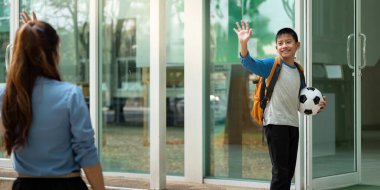 Neşeli bir çocuk annesine el sallıyor, elinde futbol topu ve sırt çantasıyla, okuldan eve dönerken heyecan ve bağımsızlığı simgeliyor..