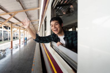 Neşeli genç bir adam, yolculuğunda önünde uzanan maceralar için heyecanla, tren penceresinden el sallıyor..
