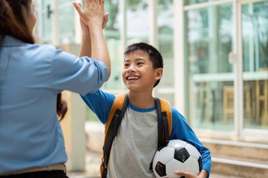 Neşeli bir çocuk, okula dönüp futbol oynayacağı için heyecanlı bir şekilde öğretmenine beşlik çakıyor. Bu, öğrenme ve spor yapma hevesini yansıtıyor..