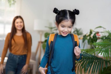 Neşeli genç bir kız okula gitmek için kapıya doğru yürürken parlak bir sırt çantası takıyor. Gülümseyen annesi, aile desteğinin sıcaklığını vurguluyor..