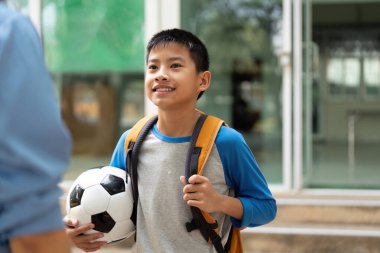 Parlak bir gülümsemeyle, genç bir çocuk okuldan sonra annesiyle konuşurken gururla futbol topunu tutuyor, hikayelerini ve kahkahalarını paylaşmaya hazır..