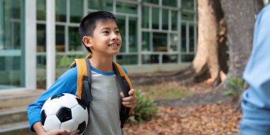 Sırt çantası ve futbol topuyla neşeli genç bir çocuk, yakınlardaki bir yetişkine gülümsüyor. Okul günü için heyecanlı. Yeni başlangıçların neşesini yansıtan, parlak ve misafirperver bir ortam..