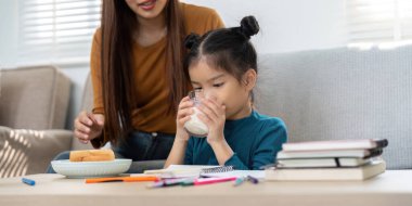 Genç bir kız süt içiyor, yanında destekleyici bir bakıcı ile ders çalışırken sağlıklı bir şeyler atıştırıyor..