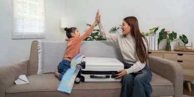 Sıcak bir oturma odasında, bir anne ve kız eğlenceli bir anın tadını çıkarırlar, bavullarının yanında otururken seyahat planlarına beşlik çakarlar..