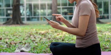 Odaklanmış bir kadın huzur parkındaki bir paspasın üzerinde oturmuş, akıllı telefonundaki sağIık uygulamalarını keşfediyor ve sağlık alışkanlığını geliştiriyor..