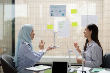 İyi aydınlatılmış bir ofiste, iki kadın ESG çerçevelerini tartışıyor ve yenilenebilir enerji girişimleri hakkında görüşlerini paylaşıyor ve örgütleri içinde sürdürülebilirlik kültürünü teşvik ediyorlar..