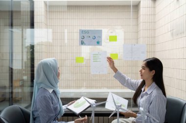 Cam duvarlı bir ofiste, iki kadın ESG girişimleri hakkında dinamik bir tartışmaya girişiyor, sürdürülebilir uygulamaları geliştirmek için verileri analiz ediyor ve içgörüleri paylaşıyor..