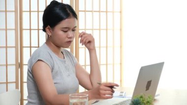 Evde oturan Asyalı bir kadın bilgisayarını kullanarak gelirini ve harcamalarını kaydediyor. Düzenli ve finansal olarak bilinçli kalıyor..