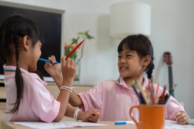 İki genç arkadaş, bir okul ortamında çocukluğun neşe ve spontanlığını vurgulayan renkli kalemler kullanırken eğlenceli bir etkileşimi paylaşıyorlar..