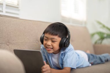 Bir çocuk kanepede yatıyor, tabletine dalmış, eğlenceli bir öğrenme deneyiminin tadını çıkarıyor. Bu resim, teknolojinin eğitimle bütünleşmesini vurgular.