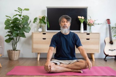 Huzurlu bir ev ortamında meditasyon yapan yaşlı bir adam yoga yaparken sağlığına, zihinsel huzura ve rahatlamaya odaklanıyor..