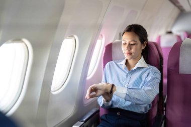 Genç bir kadın, iş seyahati süresince zaman bilincini temsil eden bir uçakta otururken saatini incelikle kontrol ediyor..