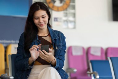 Kot ceketli genç bir kadın havaalanındaki bir salonda uçuşunu beklerken akıllı telefonuyla mutlu mesut meşgul olarak seyahat beklentisini gösteriyor..