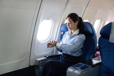 Profesyonel bir kadın uçak penceresinin yanında otururken saatine bakar, yolculuk sırasında zaman yönetimini dengeler ve seyahat eder..