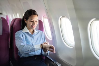 Gülümseyen bir iş kadını, uçakta otururken saatini kontrol ediyor, yolculuğunda profesyonelliği rahatlıkla harmanlıyor..
