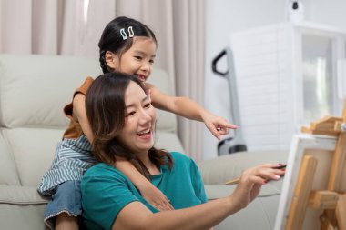 Neşeli bir kız, evde yaratıcı bir sanat oturumu sırasında annelerinin arkasında otururken tabloyu işaret eder..