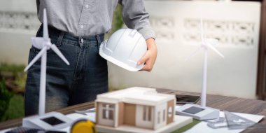 Miğfer tutan bir mühendis rüzgar türbinlerinin yer aldığı sürdürülebilir mimari bir modeli inceliyor, çevre dostu bina tasarımında yenilik sergiliyor.