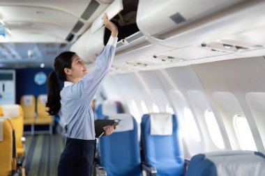 Asyalı bir kadın çantasını bir uçağın üst kompartımanına koymak için uzanıyor. Organizasyonunu ve yolculuğuna hazır olduğunu gösteriyor..