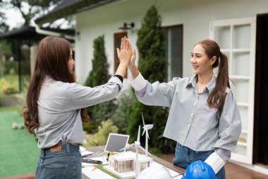 İki sürdürülebilirlik mühendisi başarılı projelerini kutlamak için açık havada bir beşlik çakıyor ve planlar ve modeller masalarında sergileniyor..