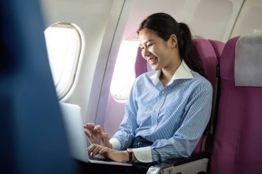 Neşeli genç bir kadın bir uçakta dizüstü bilgisayarıyla meşgul oluyor, işle bağlantılıyken iş seyahatinin tadını çıkarıyor..
