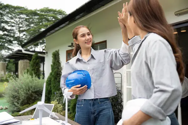 Açık havada beşlik çakan iki sürdürülebilirlik mühendisi modern bir binanın önünde proje başarılarını modeller ve planlarla kutluyorlar..