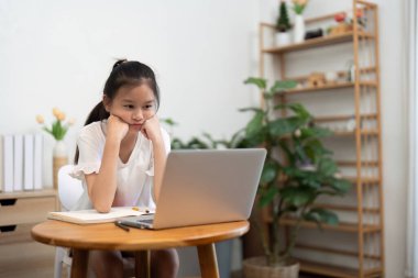 İnternette çalışırken masasına yaslanan odaklanmış bir kız, bilgisayarına dikkatlice bakıyor ve yanında bir not defteri var..