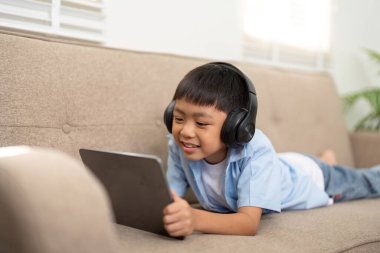 Neşeli bir çocuk kanepeye uzanıyor, kulaklık takıyor ve okula dönüş ruhunu somutlaştıran bir tabletle meşgul oluyor..