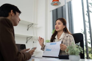 Genç bir kadın muhasebeci, erkek müşterisiyle finansal verileri tartışıyor. Modern ofis ortamında çizelgeler ve raporlar sergiliyor..